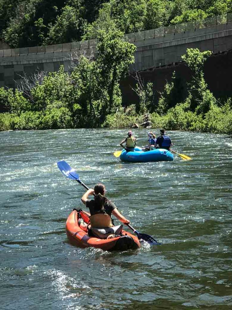 A person kayaking in a red kayak and a group rafting in a blue raft during a river trip on the Provo River.