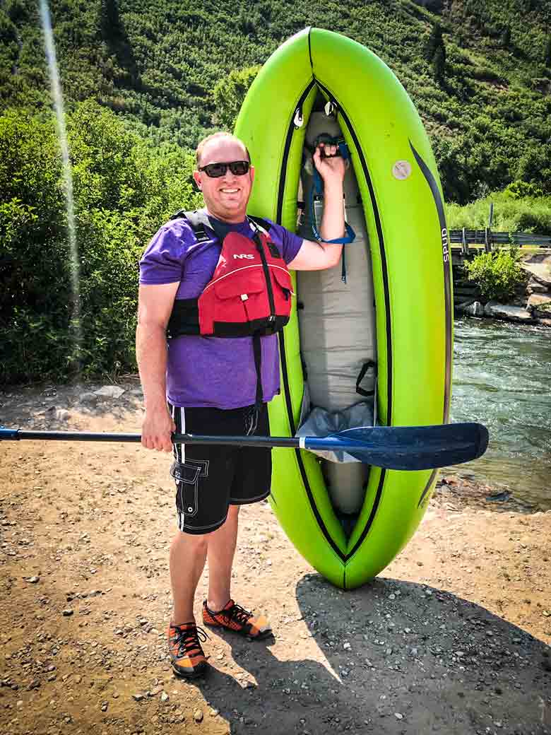 A man wearing a red life jacket standing while holding a green kayak and a blue paddle before a kayak trip on the Provo River in Utah.